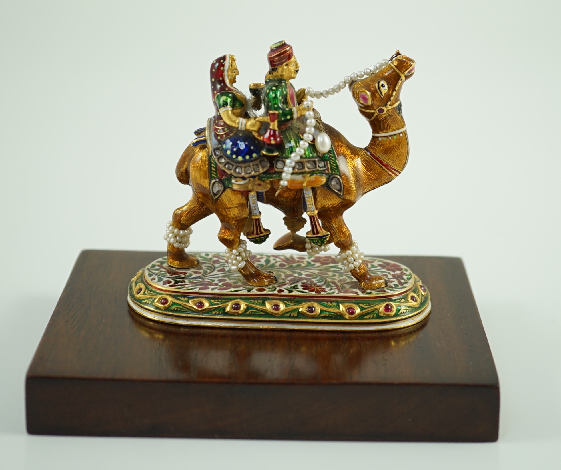 A 20th century Indian Mughal style gold, polychrome enamel, diamond, seed pearl and cabochon ruby set miniature model of a camel with two riders, on oval base